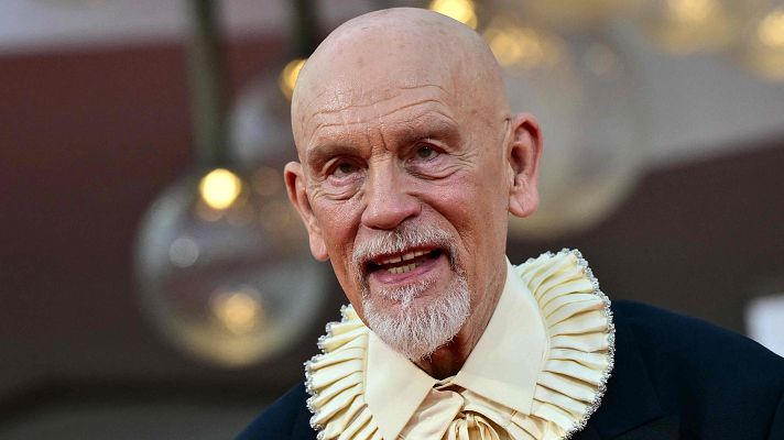 John Malkovich, hombre mayor con barba blanca y cuello de camisa voluminoso y adornado, sonríe levemente ante un fondo bokeh de luces cálidas.