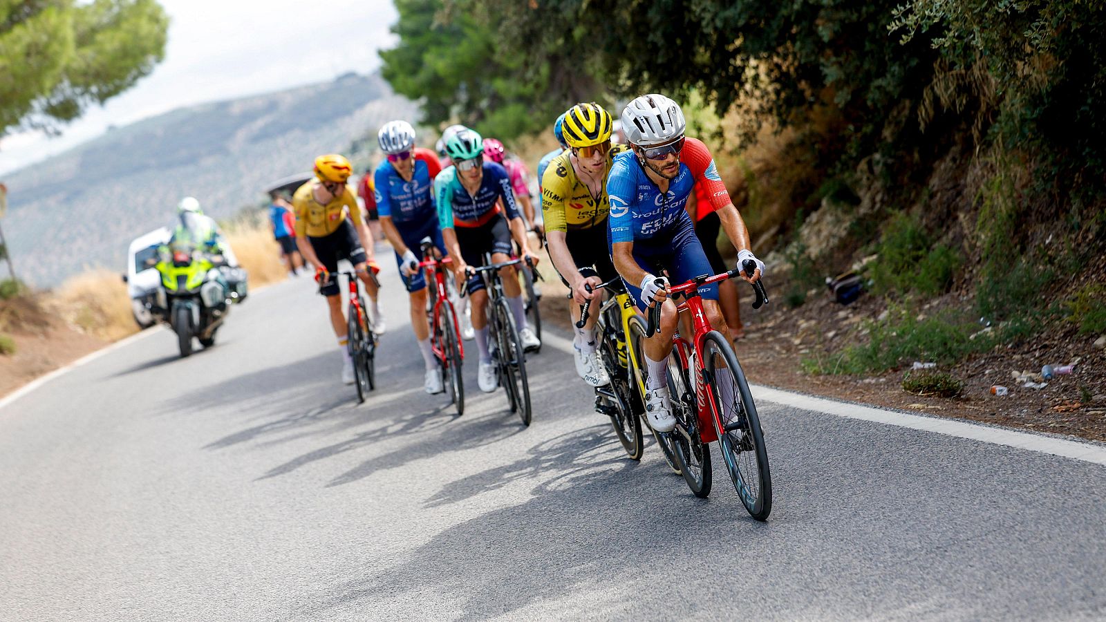 Ciclistas en plena competición en una carretera soleada y ascendente. Se observa un maillot azul liderando, seguido por uno amarillo. Una motocicleta de escolta acompaña al grupo.