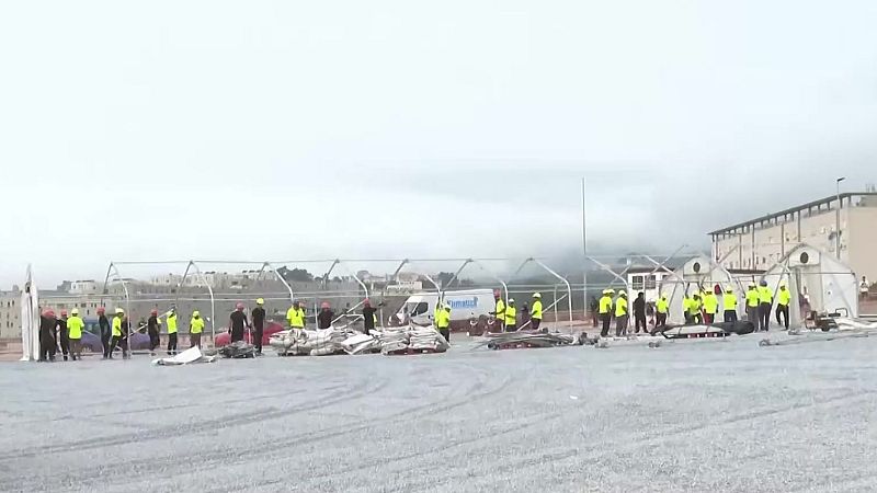 Trabajadores con chalecos de alta visibilidad y cascos montan estructuras para tiendas en un terreno al aire libre bajo un cielo nublado. Se aprecian materiales y una furgoneta blanca.