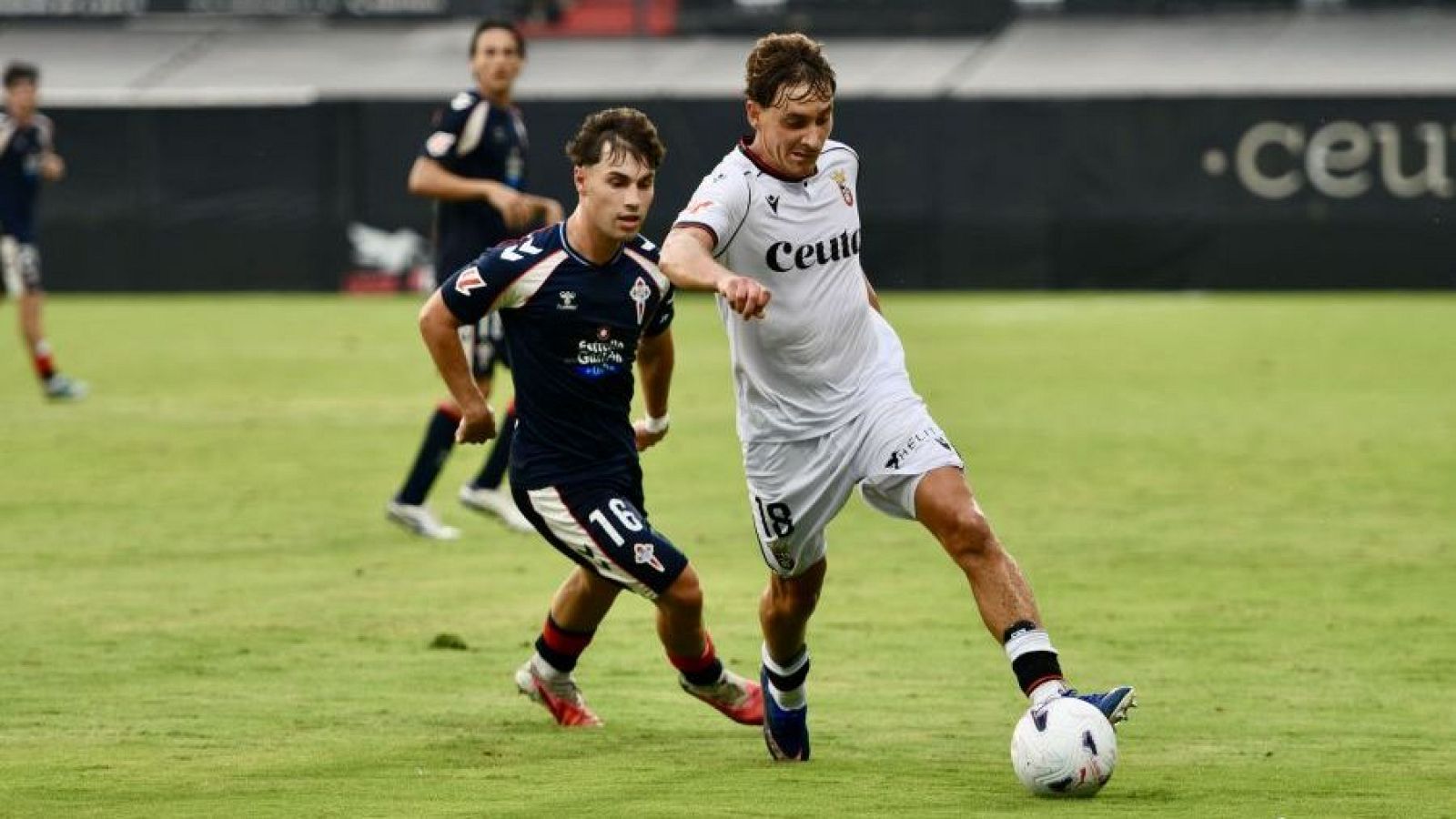 Jugador del CEUTA (dorsal 18, camiseta blanca) dribla balón ante jugador del Celta B (dorsal 16, camiseta azul oscura) en partido de fútbol.