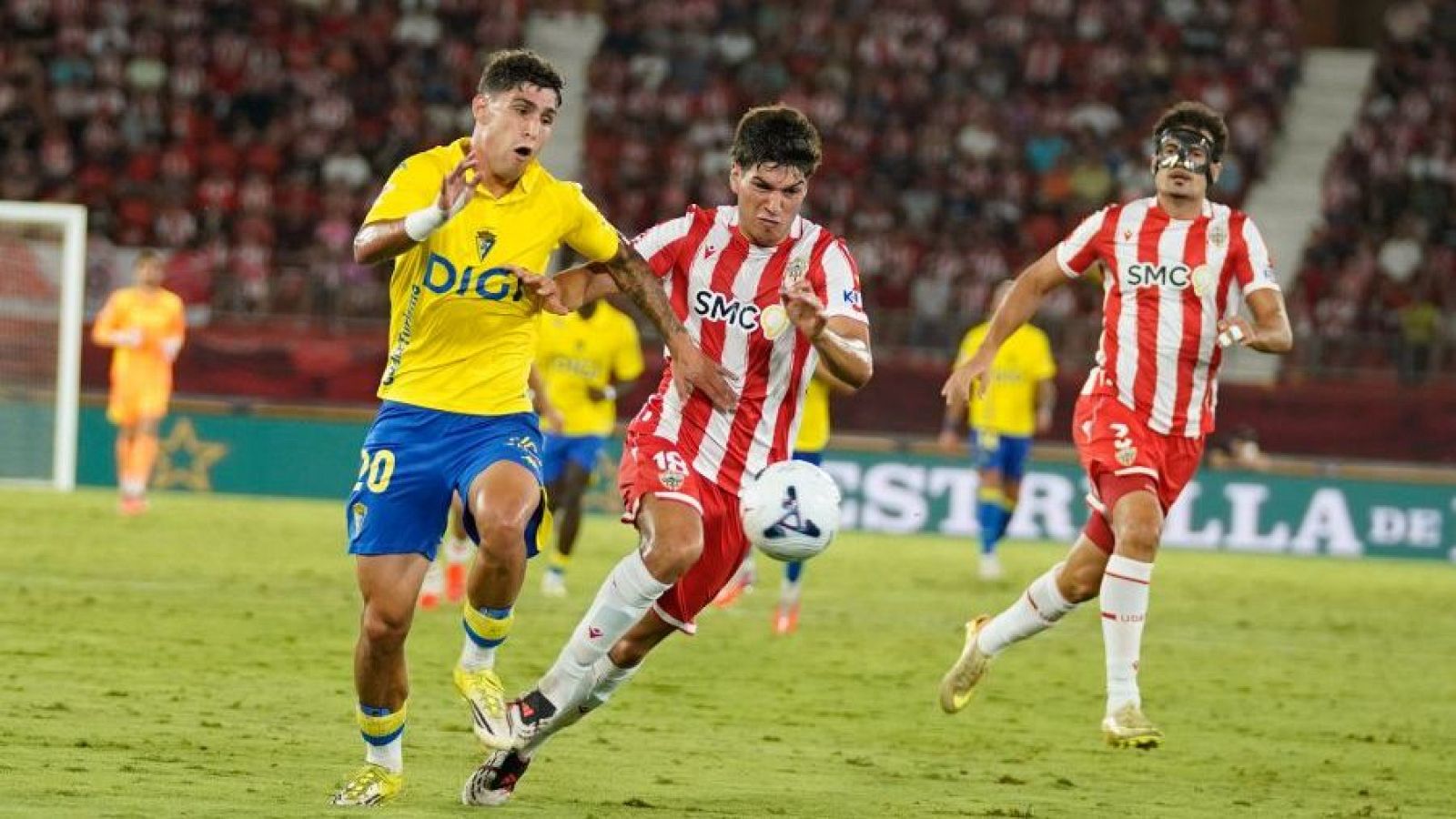 Jugadores del Cádiz CF (dorsal 20) y UD Almería (dorsales 18 y 3 con máscara) en acción durante un partido de fútbol nocturno. El balón está en juego en un estadio lleno.