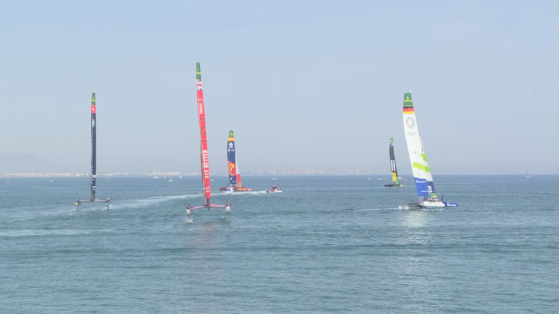 Catamaranes F50 del SailGP compiten en Valencia. Destaca el barco número 21 con bandera alemana y logo olímpico. Se lee "FLY BETTER" en otro barco.