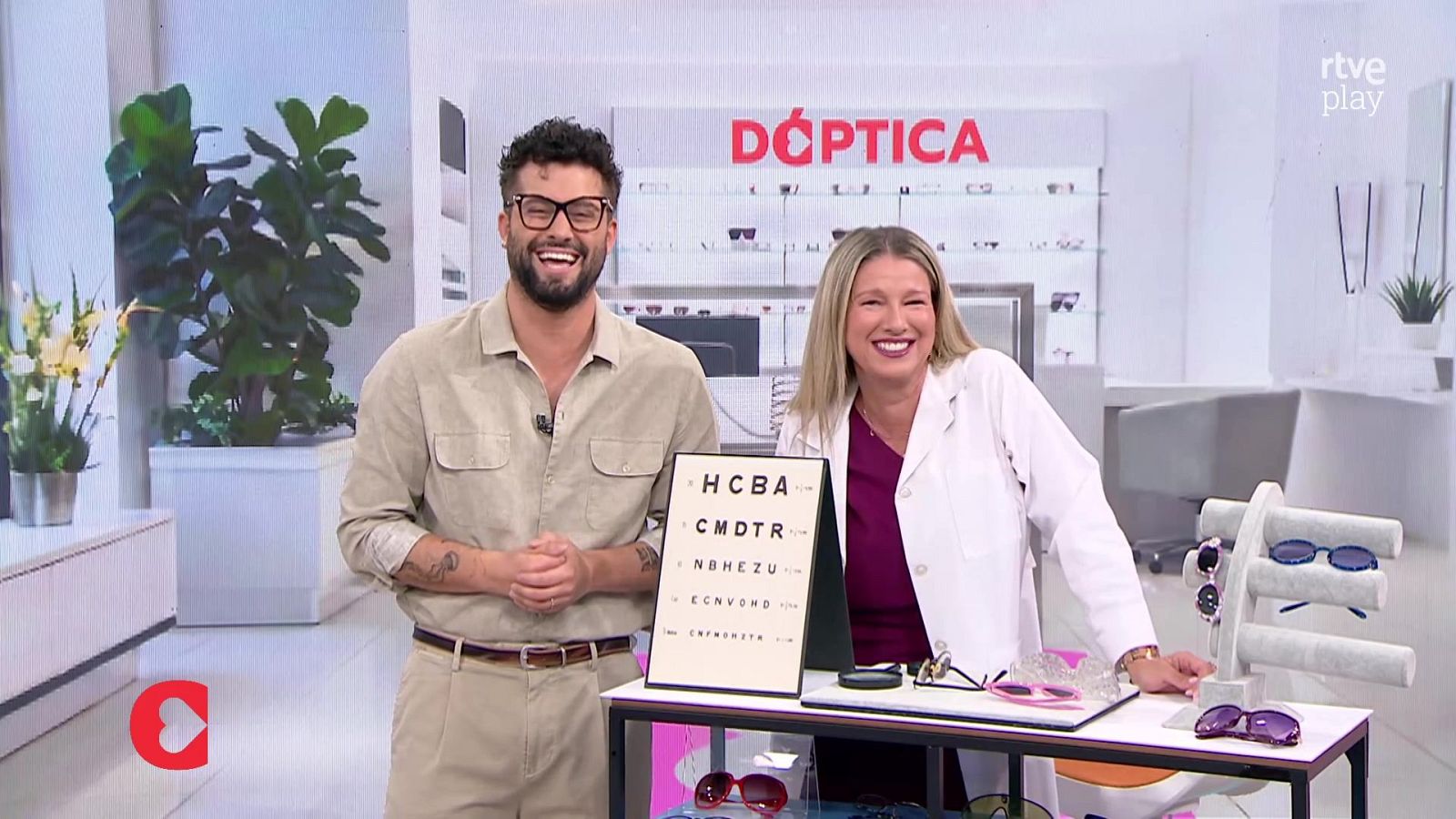 Jordi Cruz y una experta en óptica sonríen en un plató temático. Se aprecian optotipos y gafas, con el logo de rtve play y Corazón.