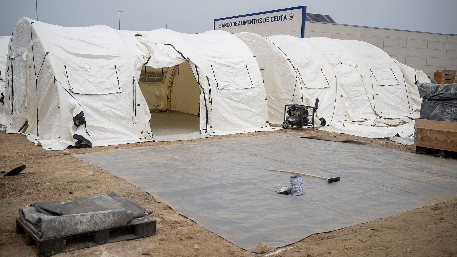 Tiendas blancas modulares y un generador eléctrico frente al "BANCO DE ALIMENTOS DE CEUTA". En primer plano, lona gris con herramientas y materiales. Cielo nublado.