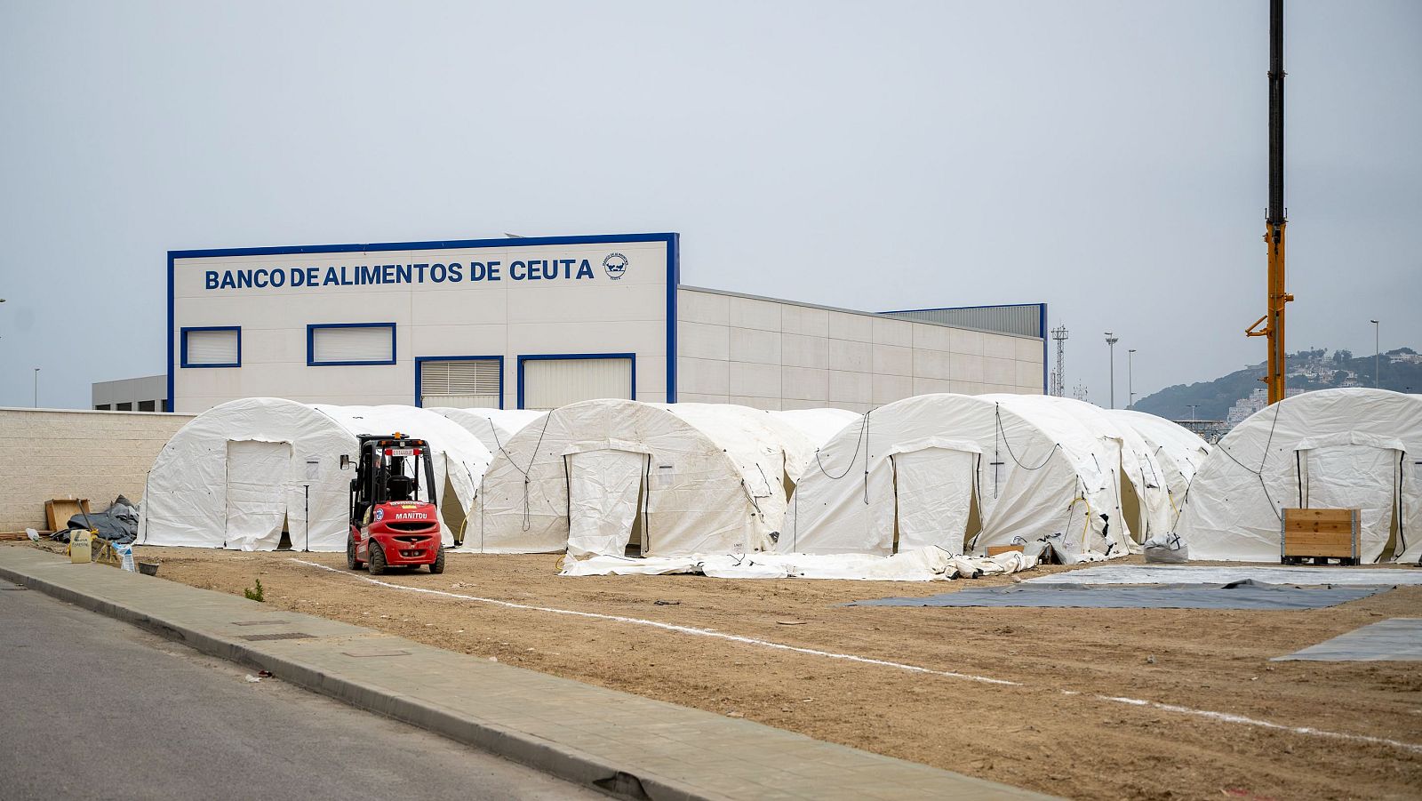 Vista exterior del Banco de Alimentos de Ceuta con varias tiendas de campaña y un montacargas Manitou. Al fondo, colinas y una grúa de construcción.