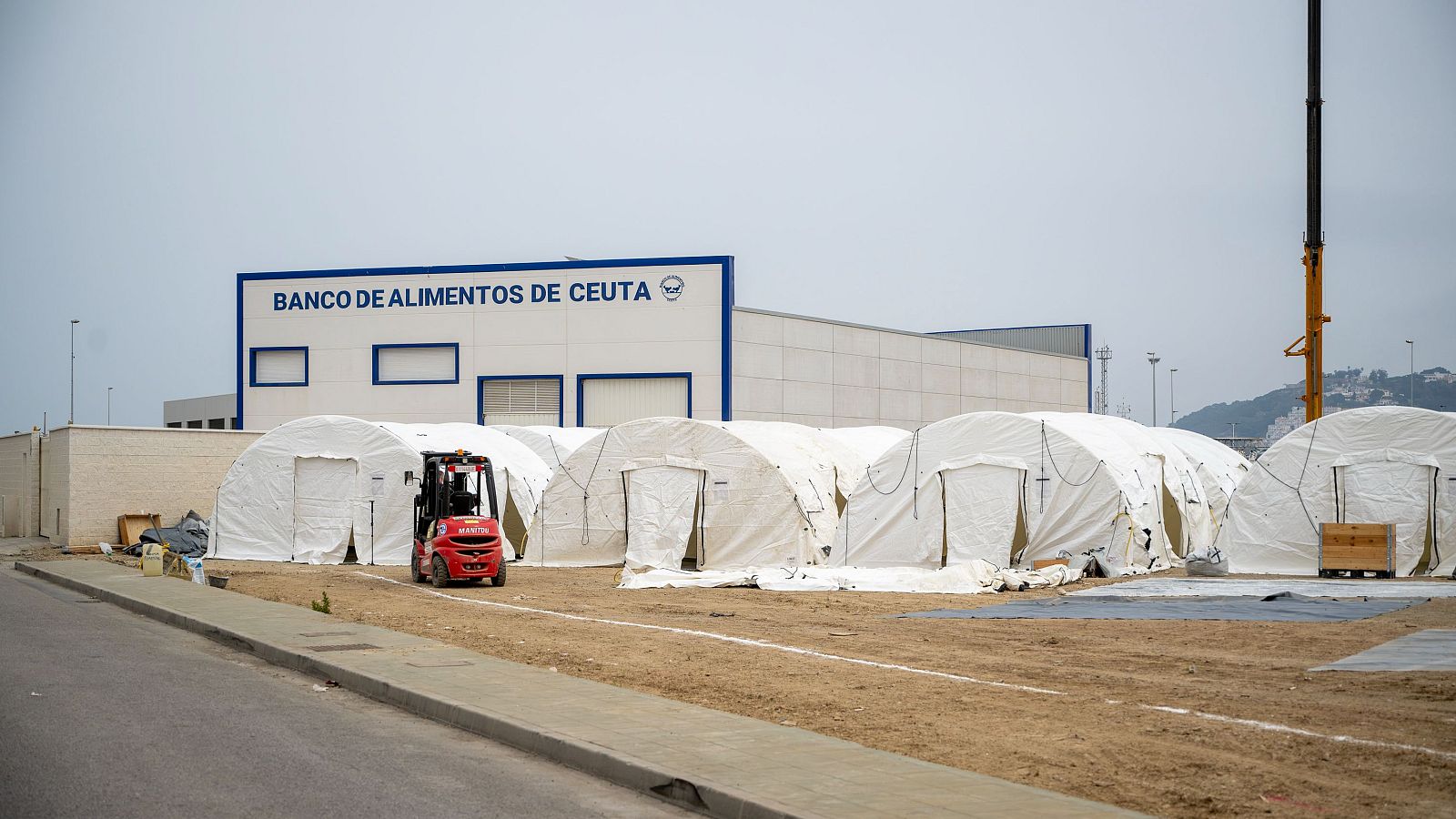 Vista exterior de un banco de alimentos con tiendas de campaña, montacargas MANITOU y grúa, bajo un cielo nublado.