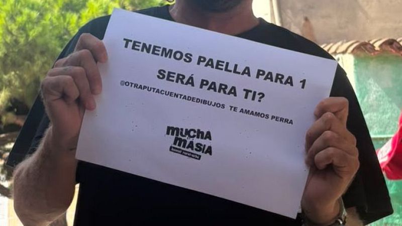 Persona con camiseta oscura sostiene cartel con mensaje sobre paella y dedicatoria a cuenta de redes sociales, logo "muchA MÁSIA" y "hotel rural-urba".