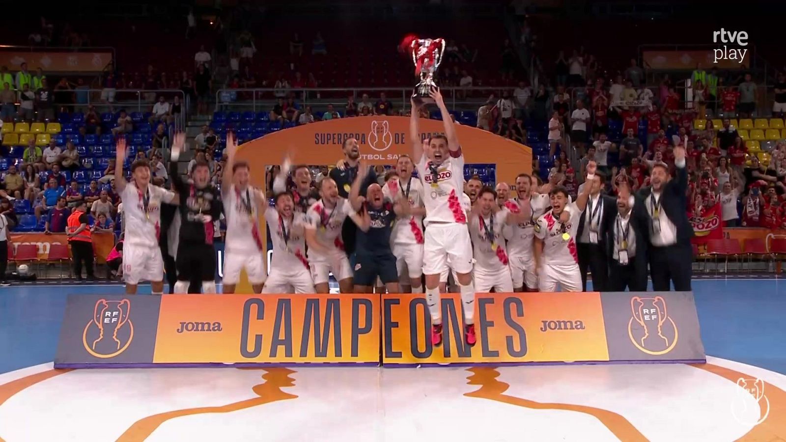 Jugadores de fútbol sala celebran la "SUPERCOPA DE FÚTBOL SALA" con un trofeo y medallas, mientras un banner anuncia "CAMPEONES". El evento es transmitido por "rtve play".