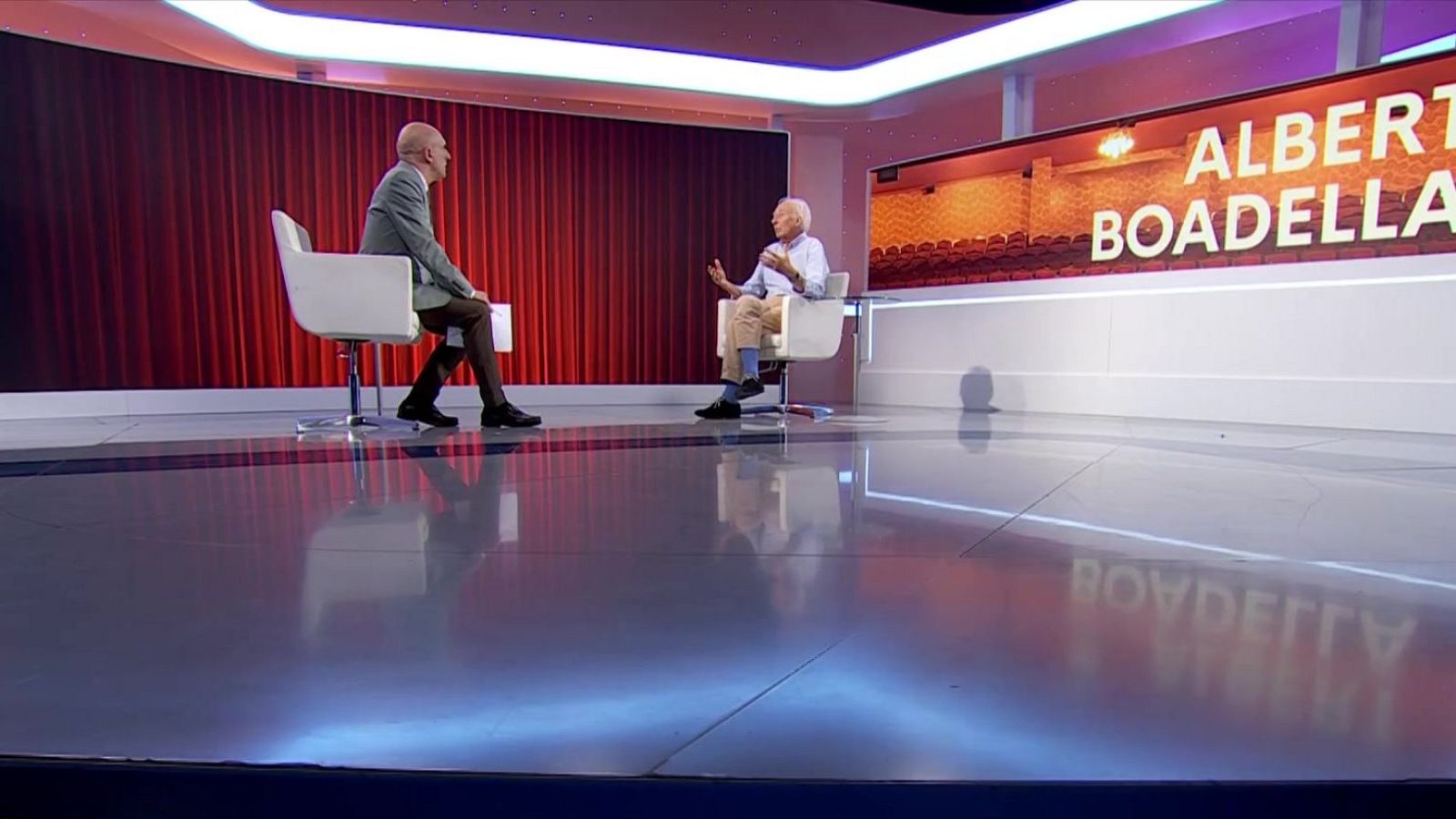 Albert Boadella, vestido con camisa azul claro y pantalones beige, gesticula mientras es entrevistado en un plató con pantalla que muestra su nombre y un auditorio.