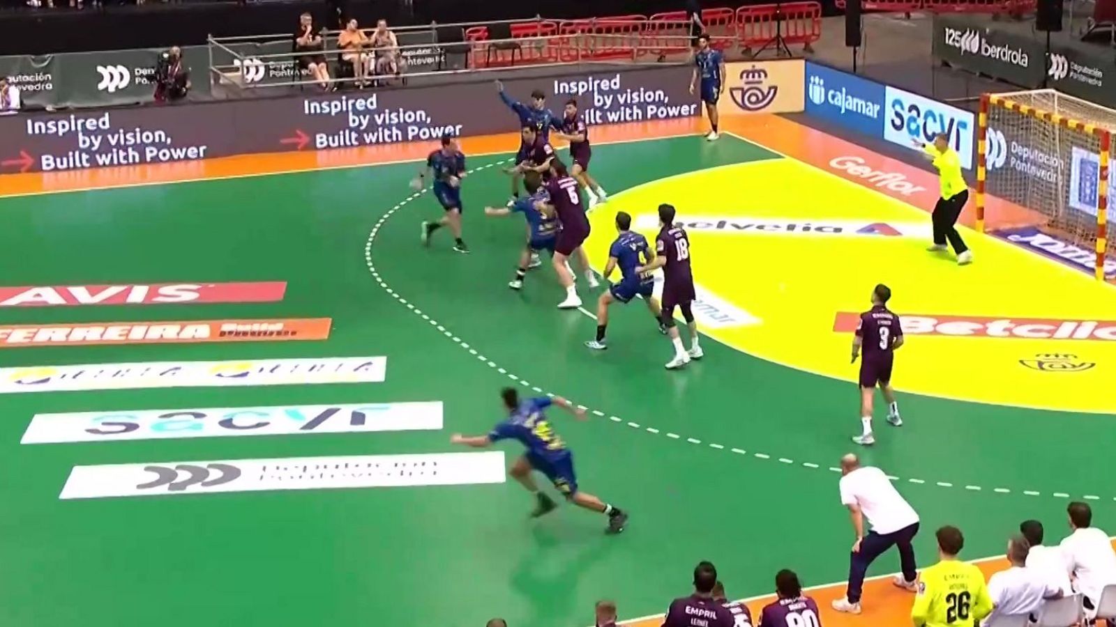 Vista aérea de un partido de balonmano con jugadores de equipos azul/morado y amarillo/negro. Se aprecian números de jugadores, publicidad en cancha y vallas, y gradas al fondo.