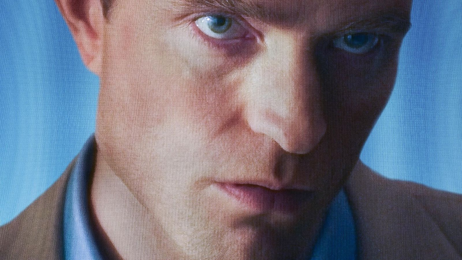 Primer plano de Benedict Cumberbatch con mirada intensa. Ojos azules penetrantes, cejas fruncidas, camisa azul y chaqueta beige. Fondo azul pixelado.