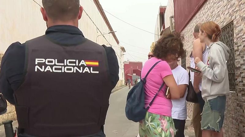Agente de la Policía Nacional en Ceuta, de espaldas, observa una calle. Civiles en segundo plano, uno con camiseta rosa y falda floral, otro con sudadera gris y falda vaquera.