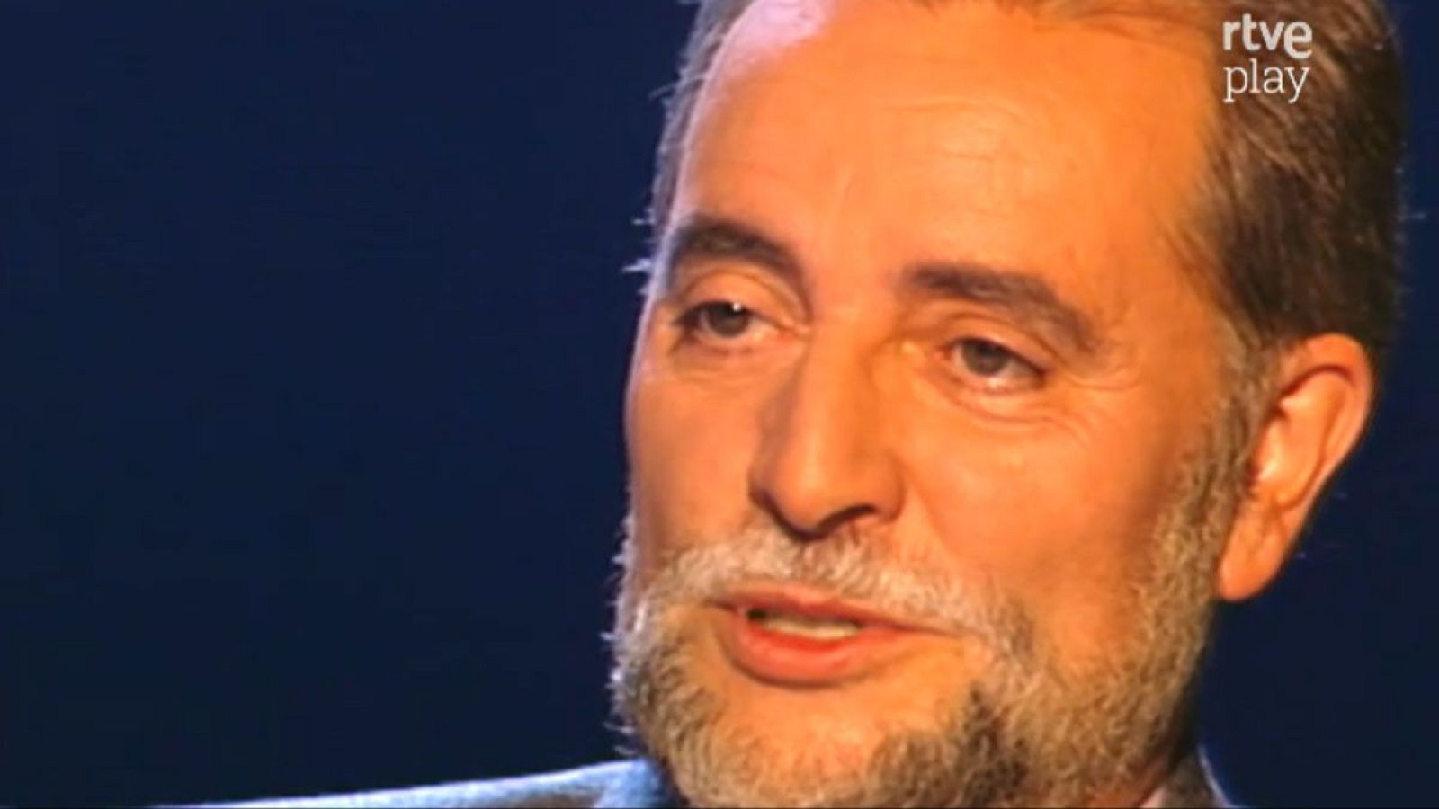 Julio Anguita, político español, en un estudio de televisión con el logo de "rtve play". Su rostro serio y barba grisácea son prominentes.