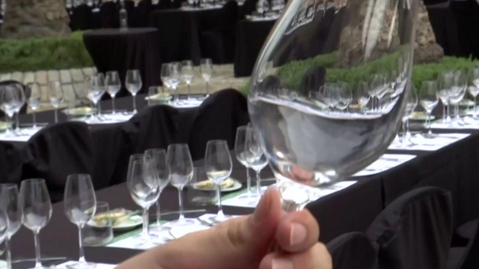 Mano sosteniendo copa de vino con líquido claro. Mesas con copas vacías y platos blancos al fondo, en un entorno exterior con vegetación.