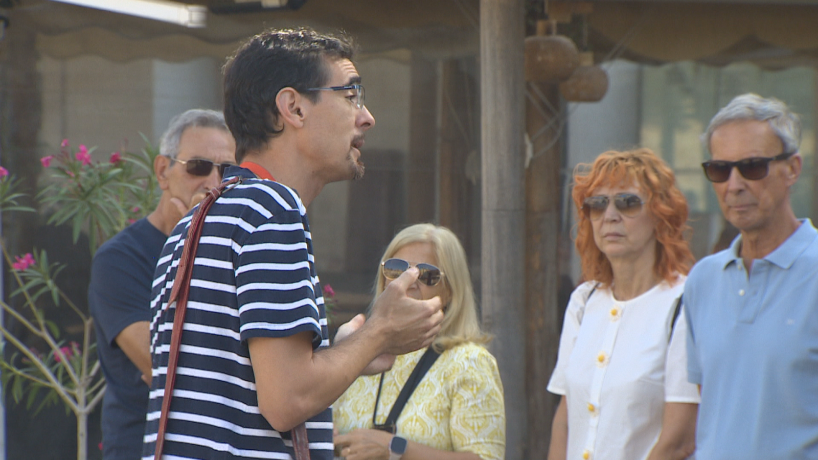 Un hombre con camiseta de rayas habla animadamente a un grupo de cuatro personas en un exterior soleado. Se aprecian detalles de vestimenta y gestos en los asistentes.