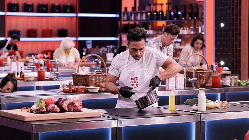 David Bustamante, con delantal de MasterChef, manipula una olla en una cocina profesional. Ingredientes como carne, lechuga y tomate a su lado. Cocina moderna con otros concursantes al fondo.