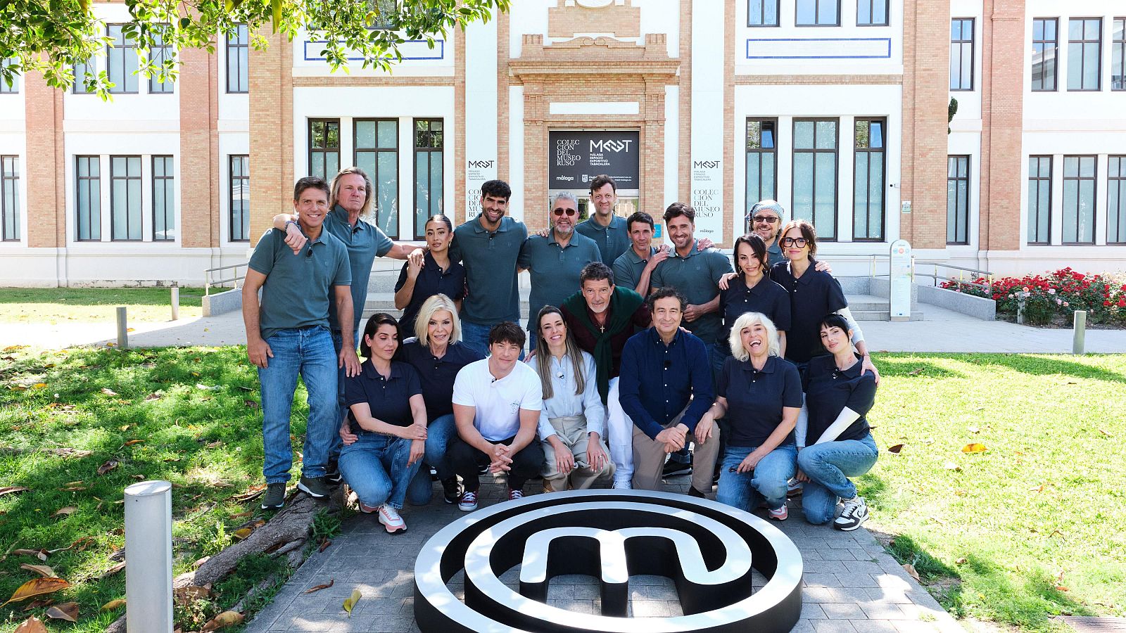 Grupo de 18 personas, entre ellas Antonio Banderas, posan frente a un edificio con logo "MOST" o "MCT". En primer plano, logo 3D de MasterChef. Visten polos azules/verdes y vaqueros.