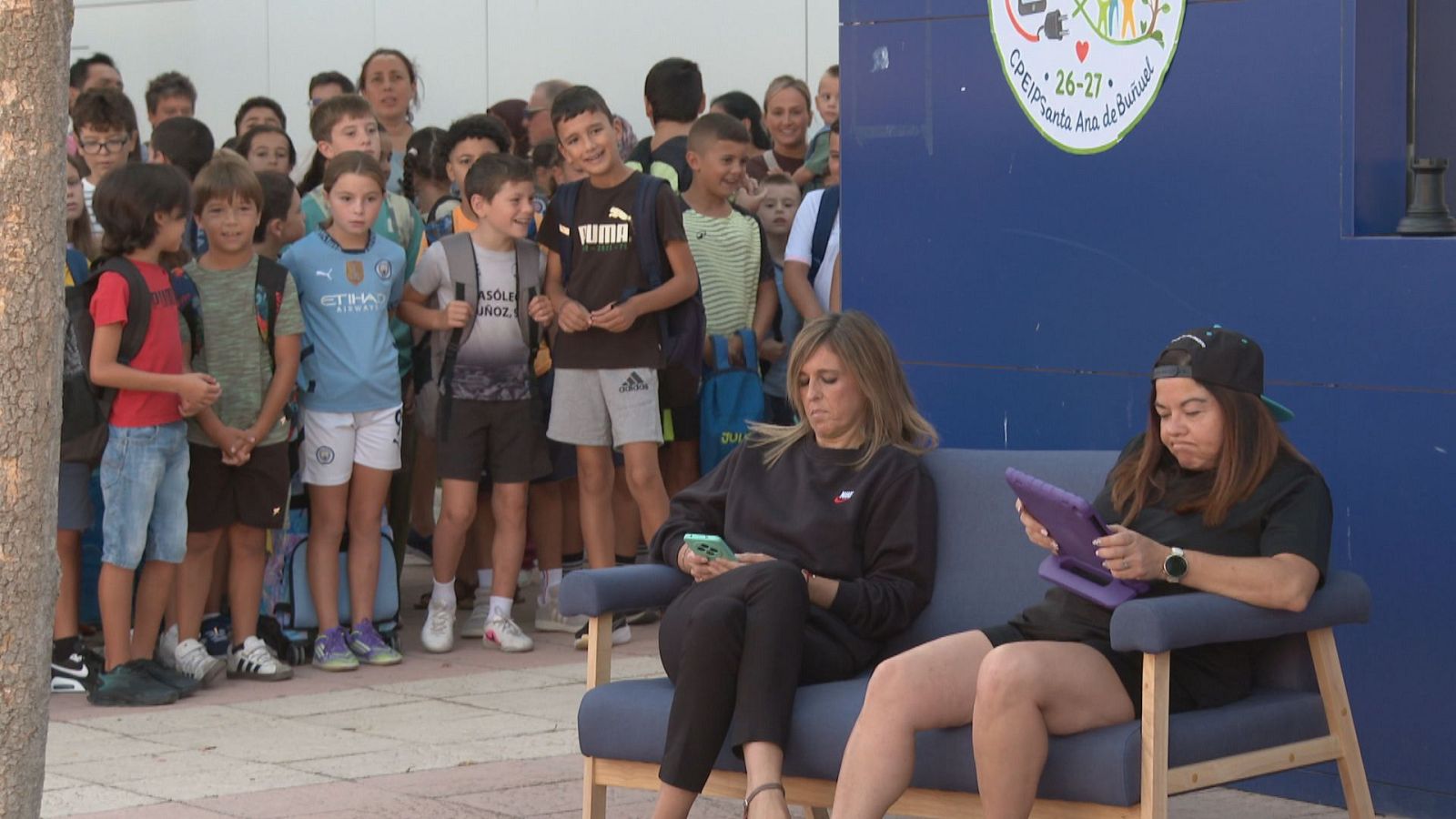 Mujeres en sofá con móvil y tableta, rodeadas de niños curiosos con camisetas de fútbol. Fondo de edificio con logo de colegio 'CPEIP Santa Ana de Buñuel'.