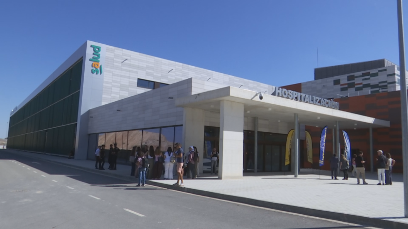 Fachada de hospital moderno con "HOSPITALIZACIÓN" visible. Grupos de personas en la acera sugieren un evento.