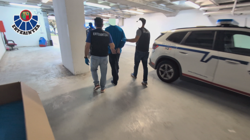 Agentes de la Ertzaintza trasladan a un detenido esposado, con chalecos identificativos y un vehículo policial a la derecha. Se aprecian el escudo de la Ertzaintza y el número de emergencias 112.