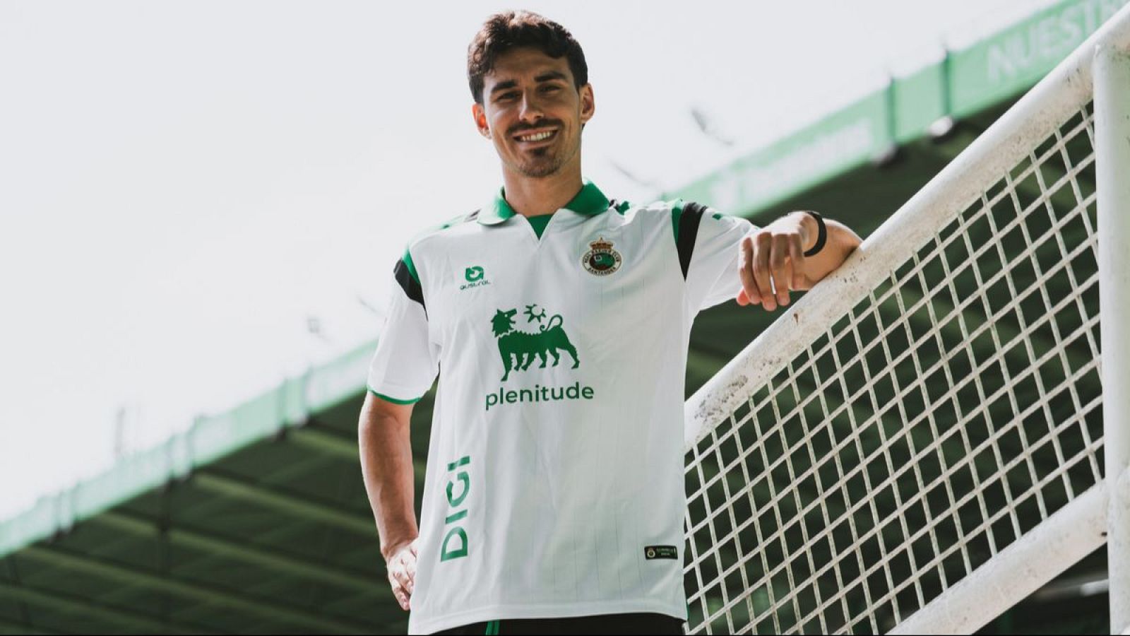 Íñigo Vicente, jugador del Racing de Santander, sonríe a cámara con la equipación del equipo, luciendo logos de Givova, Eni (plenitude) y DIGI. Fondo de estadio.