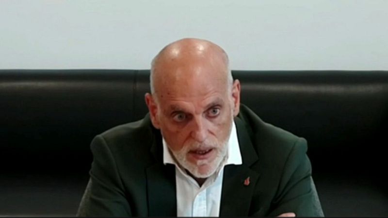 Javier Tebas, calvo y con barba blanca, mira intensamente a la cámara con una chaqueta verde oscuro, camisa blanca y un pin rojo en la solapa. Fondo dividido en oscuro y claro.