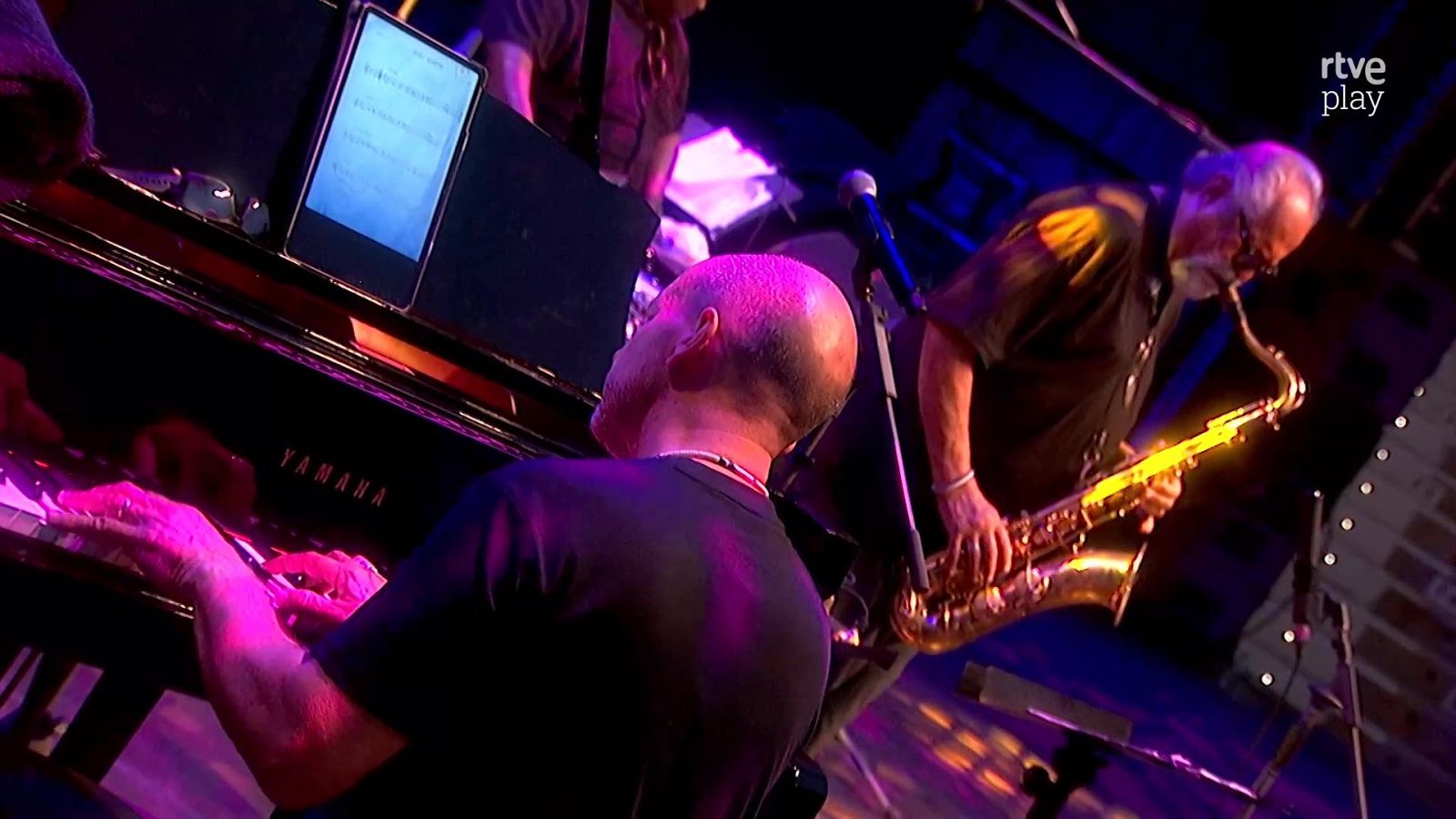 Músicos en un escenario: un pianista y un saxofonista bajo luces de colores, con un piano Yamaha y el logo de rtve play visible.