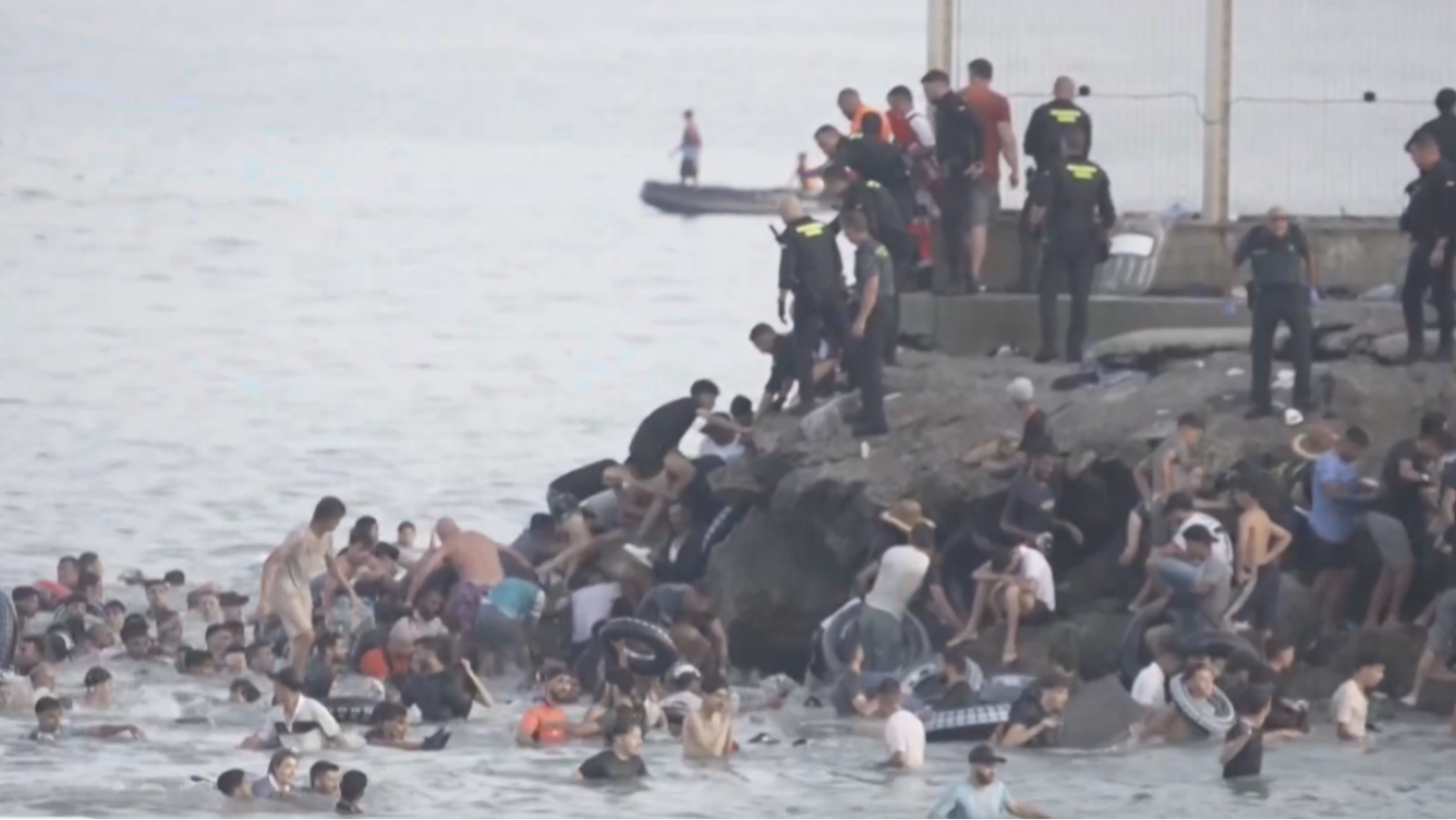 Gran afluencia de personas llegando por mar a una costa, con presencia de seguridad. Se observan rocas y una plataforma elevada.