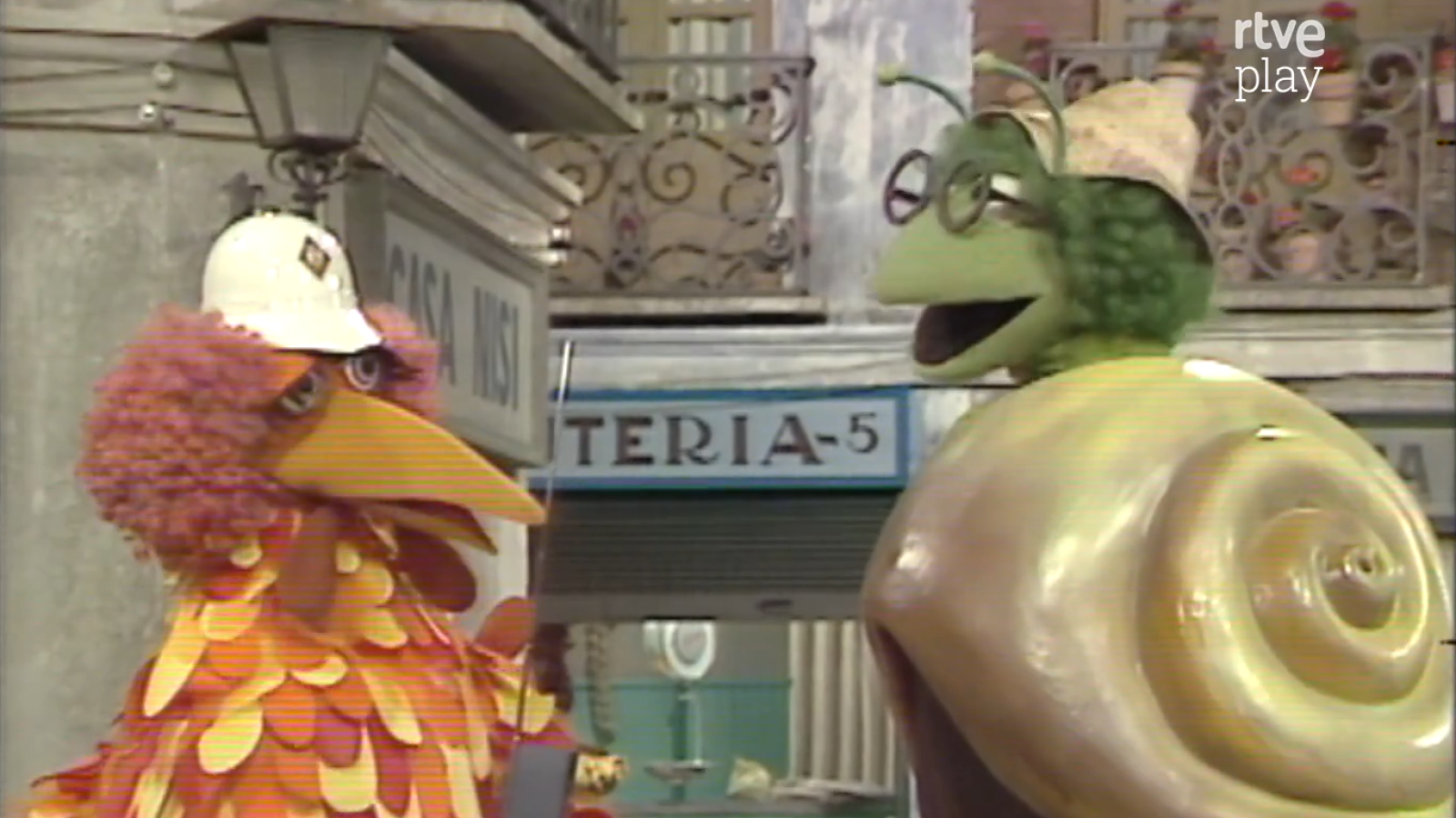 Pino, el pájaro de Barrio Sésamo, y Perezgil, el caracol con gafas, en una calle con edificios y el logo de rtve play.