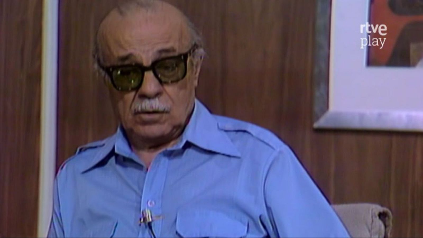 Primer plano de Ernesto Sábato, escritor argentino, con bigote blanco, gafas oscuras y camisa azul. Fondo de madera oscura, cuadro abstracto y logo de RTVE. Imagen de archivo de RTVE.