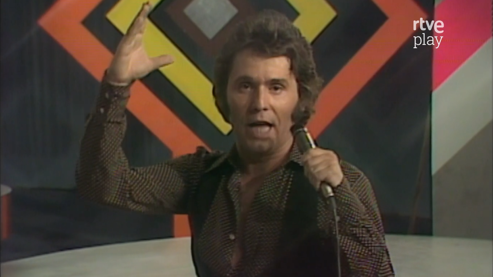 Primer plano de Raphael cantando con intensidad, micrófono en mano y brazo alzado. Fondo geométrico retro con logo "rtve play".