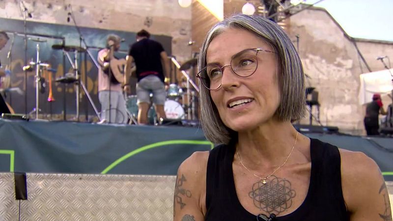 Mujer de mediana edad con pelo gris, gafas rojizas y tatuajes, sonriendo. Detrás, un escenario musical con músicos y luces.
