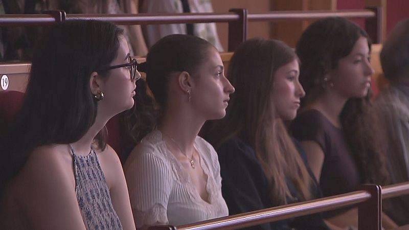 Primer plano de cuatro jóvenes mujeres sentadas en un auditorio, todas mirando hacia adelante con atención. Se aprecian detalles de sus vestimentas y peinados, en un entorno de asientos rojos y una barandilla de madera.