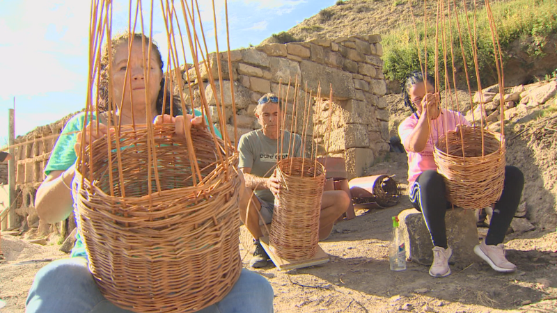 Dos mujeres y un hombre elaboran cestas de mimbre al aire libre, junto a una pared de piedra y una ladera con vegetación escasa.