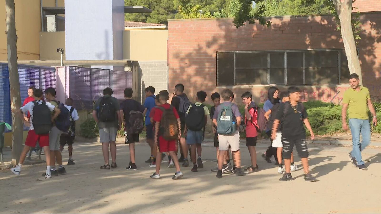 Jóvenes con mochilas en el primer día de colegio en Lleida, 8 de septiembre de 2026. Un hombre camina a la derecha. Entorno exterior con edificios y árboles.