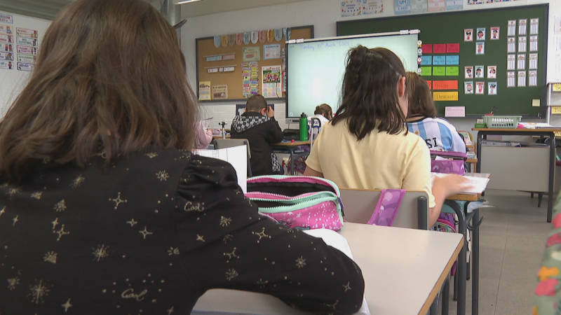 Vista de un aula escolar con alumnos y una pizarra digital que muestra "Monday 1st March". Se aprecian detalles de ropa, estuches y pizarras decoradas con días de la semana.