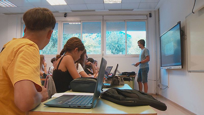 Aula moderna con estudiantes y profesor. Pupitres con portátiles, pantalla digital y ventanas con vista a árboles. El profesor, con camiseta gris y vaqueros, sostiene papeles.