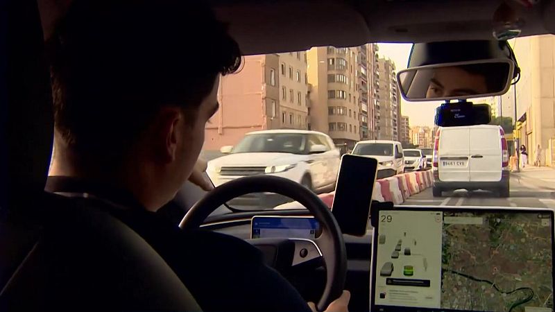 Vista desde el interior de un coche en movimiento en Zaragoza, con el conductor al volante, un móvil y una pantalla de navegación. Tráfico moderado con furgoneta blanca (8441 CYS) y barreras de obras.