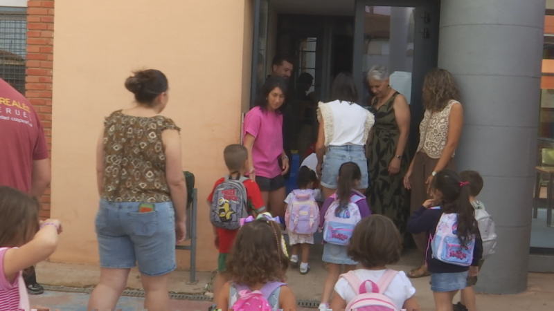 Niños con mochilas coloridas y adultos en la entrada de un edificio, listos para el inicio de la jornada escolar en Perales.