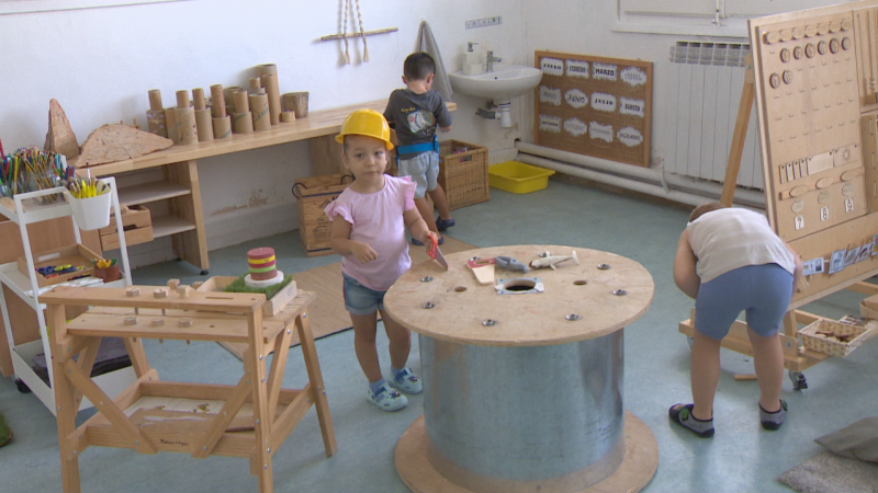 Niños en un aula de infantil jugando con un casco de juguete, en un lavabo y en un panel de actividades. Se aprecian etiquetas con partes del cuerpo en español.