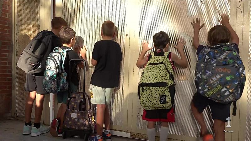 Cinco ni�os de espaldas observan curiosos a trav�s de una barrera met�lica perforada. Llevan mochilas y ropa casual, con una pared de ladrillo al fondo. Se aprecian gr�ficos de "1 Directe" en la esquina inferior derecha.