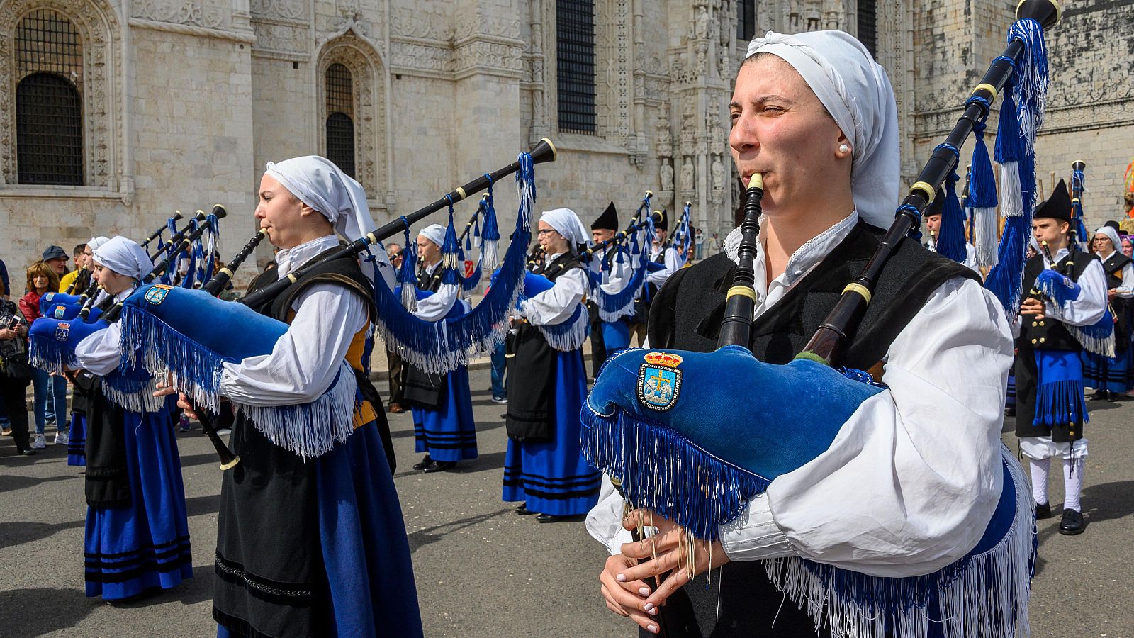 Mujer tocando una gaita azul con el escudo de Oviedo, rodeada por la Real Banda de Gaitas 'Ciudad de Oviedo' frente al Monasterio de los Jerónimos en Lisboa.