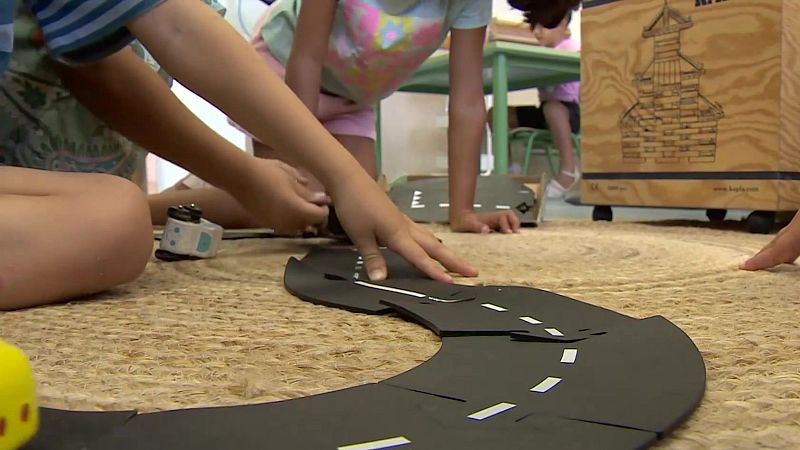 Manos de niños construyen una carretera de juguete negra sobre alfombra de yute, rodeados de otros juguetes como un robot y un objeto amarillo con puntos rojos. Al fondo, una mesa verde y una caja de madera con un dibujo oriental.