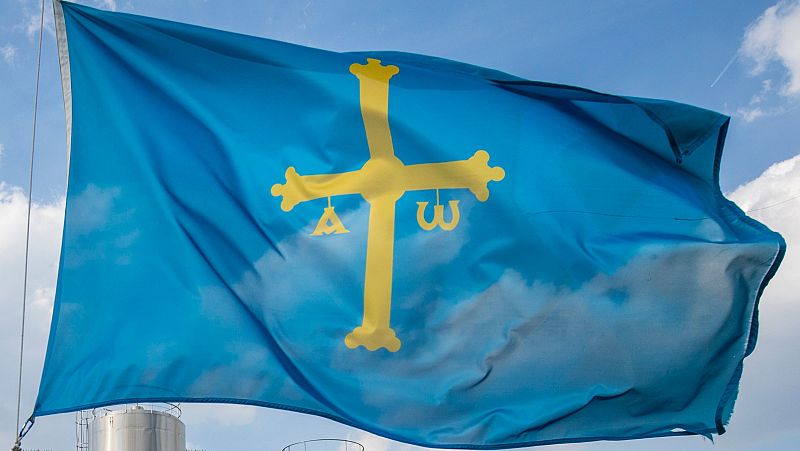 Bandera de Asturias azul con Cruz de la Victoria dorada, Alfa y Omega, ondeando bajo cielo azul con nubes. Se ve parte de un mástil y estructura metálica.