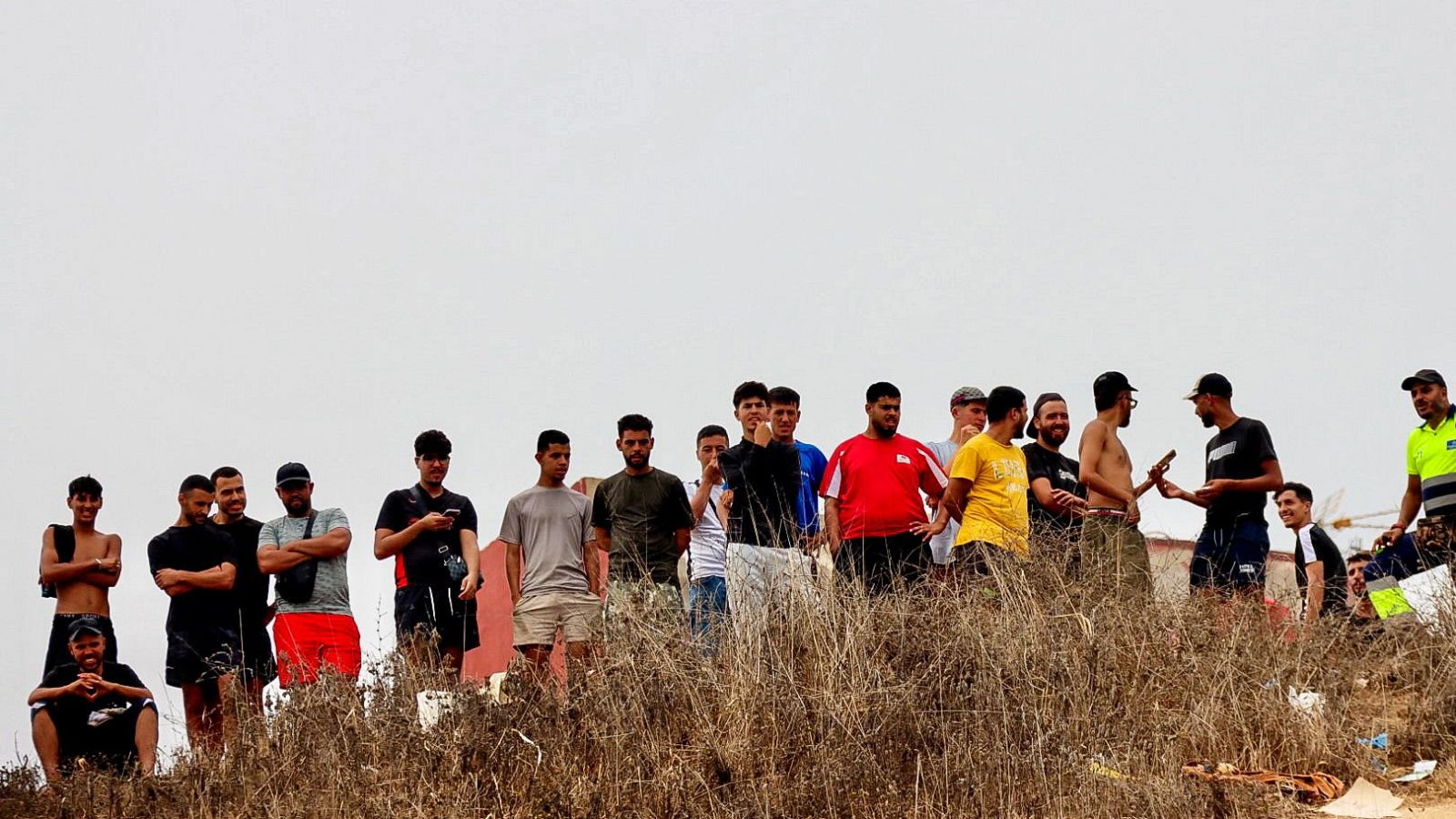 Grupo de migrantes en Ceuta, en un terreno natural con hierba seca, bajo un cielo gris. Hombres jóvenes y adultos observan o esperan, algunos con móviles.
