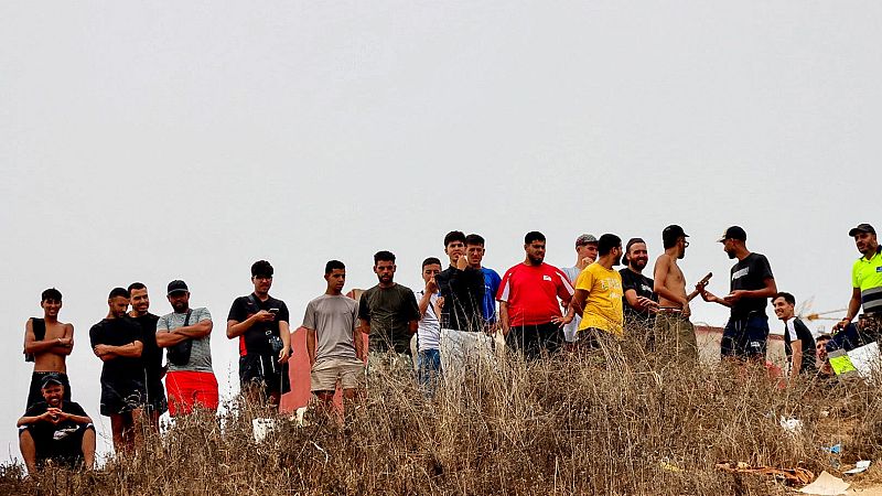 Grupo de migrantes en Ceuta, en un terreno natural con hierba seca, bajo un cielo gris. Hombres jóvenes y adultos observan o esperan, algunos con móviles.