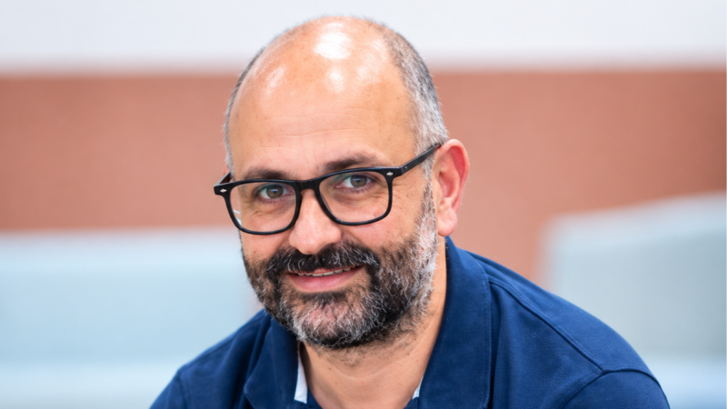 Primer plano de Andreu Buenafuente sonriendo a cámara, con gafas oscuras, barba y bigote grises, y polo azul. Fondo desenfocado con tonos melocotón y azul.