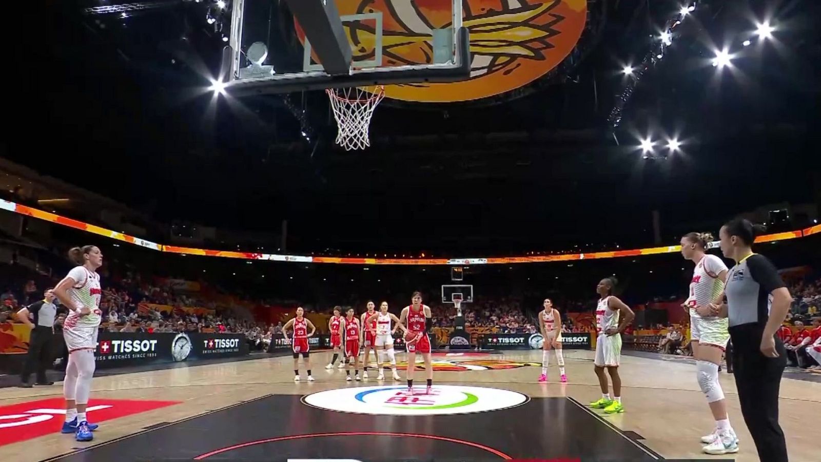 Partido de baloncesto femenino en un pabellón. Jugadoras con uniformes rojo y blanco, árbitras y paneles publicitarios de Tissot son visibles en la cancha iluminada.