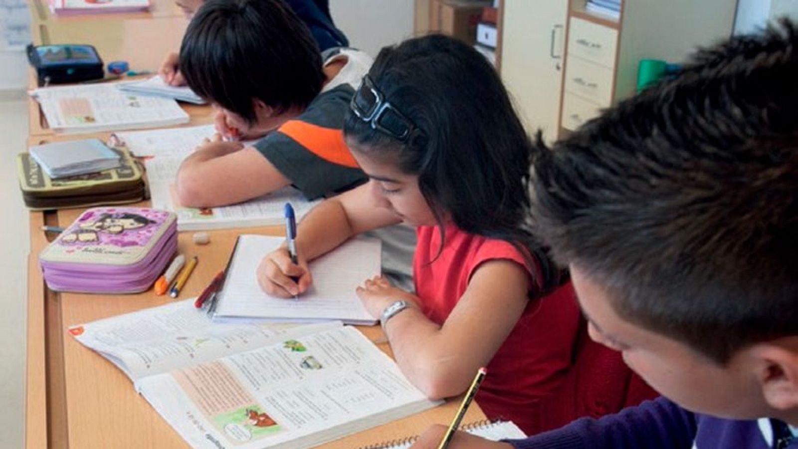Niños concentrados en sus pupitres con cuadernos, libros y material de escritura. Se aprecia un mueble de almacenamiento al fondo.