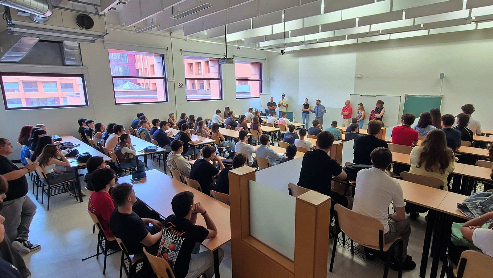 Vista de un aula universitaria con estudiantes y oradores en el frente. Se aprecian ventanas, un proyector y mobiliario de madera.